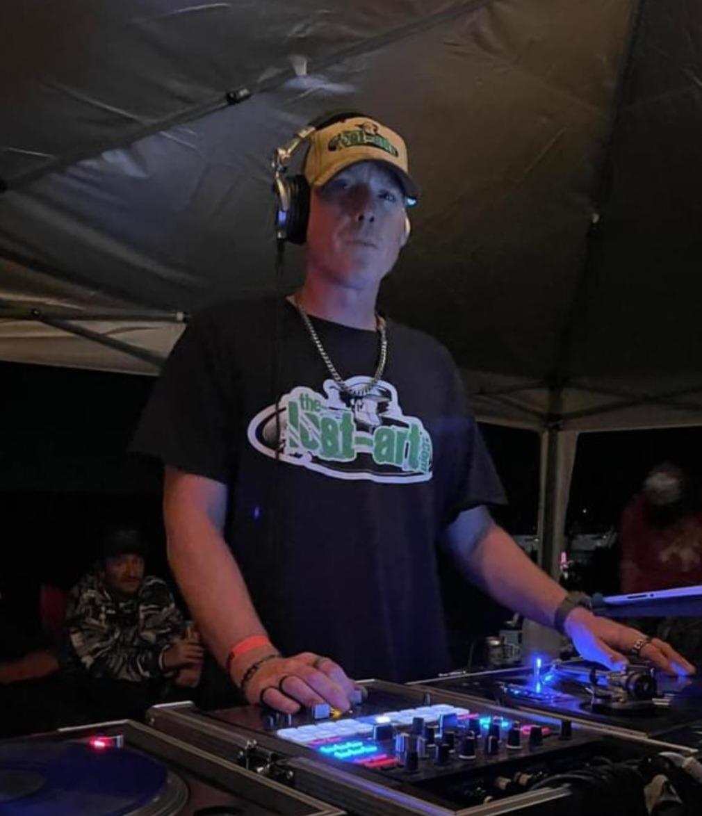 DJ Natural Nate® in The-Lost-Art™ black tee and yellow hat at outdoor tent, hands on mixer under blue LED lighting